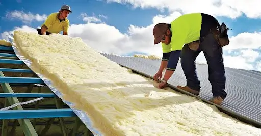 two construction workers installing a large sheet of roof insulation blanket on a metal roof
