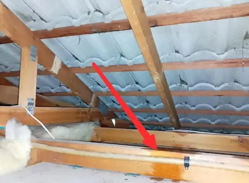 interior view of a roof cavity showing cracked grey concrete roof tiles with visible water stains on the timber framing, highlighted by a red arrow indicating a leak point.